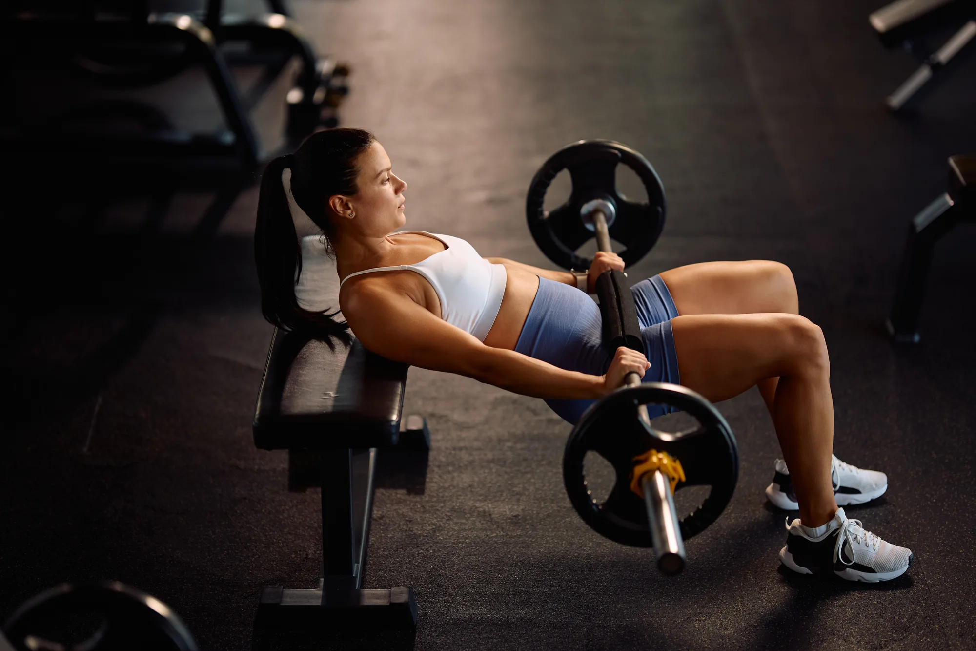 Athletic woman doing barbell hip thrust exercise while working out in a gym.