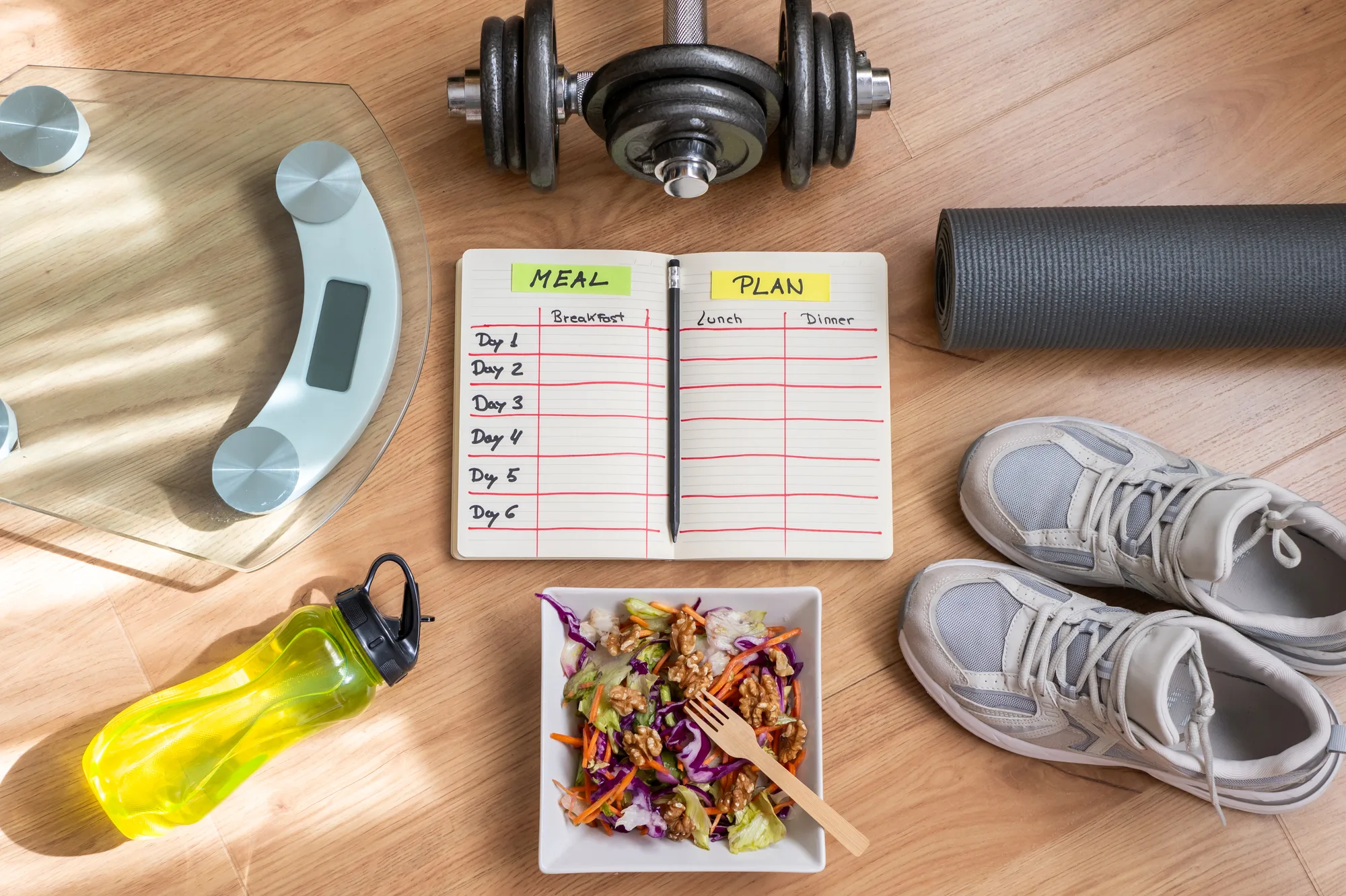 Pèse-personne, haltère, carnet de plan de repas ouvert, salade, bouteille d'eau et chaussures de sport vus de dessus sur un parquet.