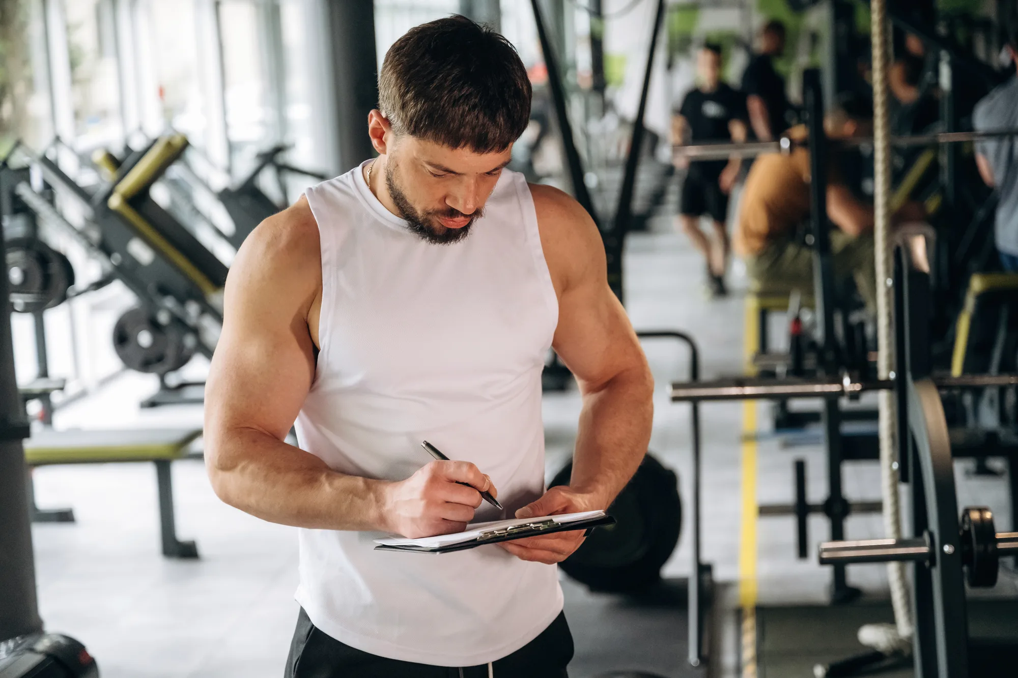 Person im weißen Tanktop schreibt in einem Fitnessstudio auf ein Klemmbrett, im Hintergrund Geräte und Racks.